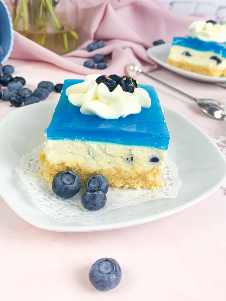 Das ist ein Bild von Ecken mit Löffelbiskuitboden, Vanillecreme. Blaubeeren und einem blauen Tortengussspiegel.
