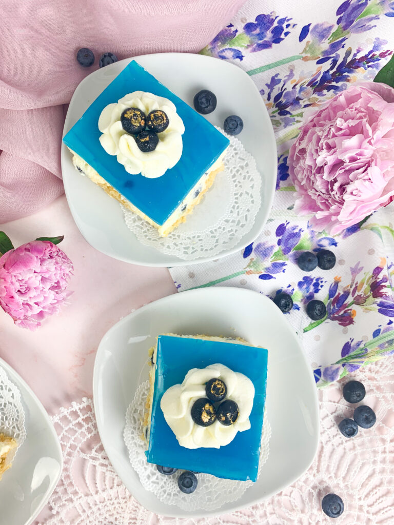 Das ist ein Bild von Blaubeer-Ecken mit Löffelbiskuitboden, Vanillecreme. Blaubeeren und einem blauen Tortengussspiegel.